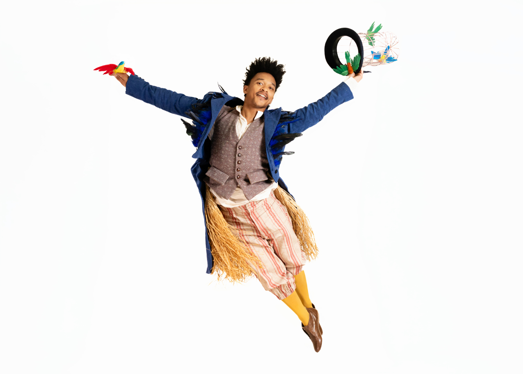 A male dancer in a long blue tailcoat, brown waistcoat, and red-and-yellow striped trousers leaps mid-air with legs together behind him. His arms stretch wide as he smiles, holding a birdcage-style top hat in one hand and a bright red-and-yellow bird in the other